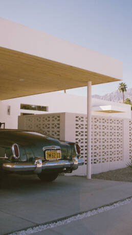 a concrete wallof breezeblocks stands in front of a home with a carport and a green vintage car in the driveway, the whole house is white, behind the home there is a palm tree, mountains, and a clear blue sky